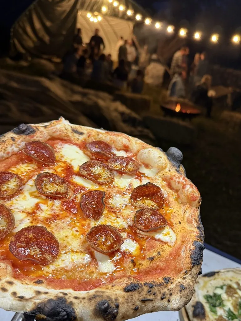 Freshly cooked pepperoni pizza held up at an evening party, with guests and festoon lights behind
