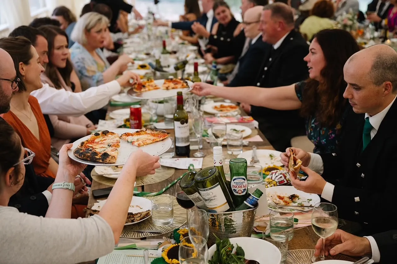 Wedding guests passing boards of pizza along a long marquee table